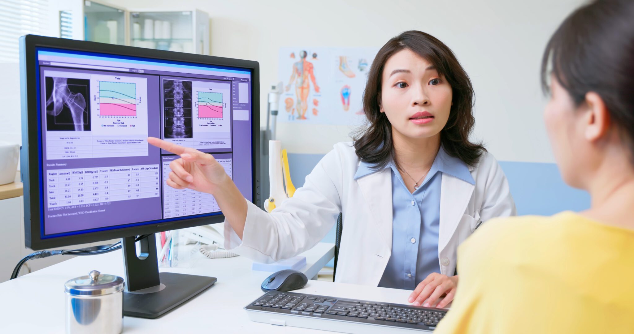 A doctor in a white coat points to a monitor displaying bone density scan results and charts while explaining them to a patient in an office.