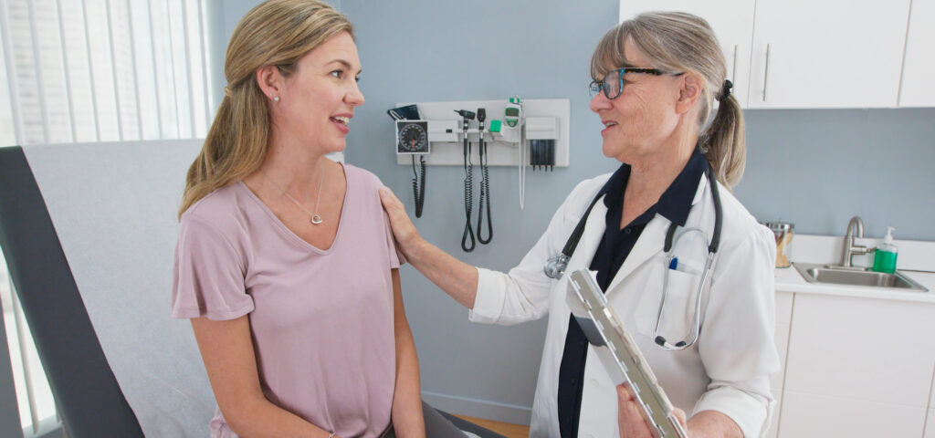 Two shot of woman talking to her reassuring primary care doctor in exam room. Middle aged patient having appointment with female senior physician