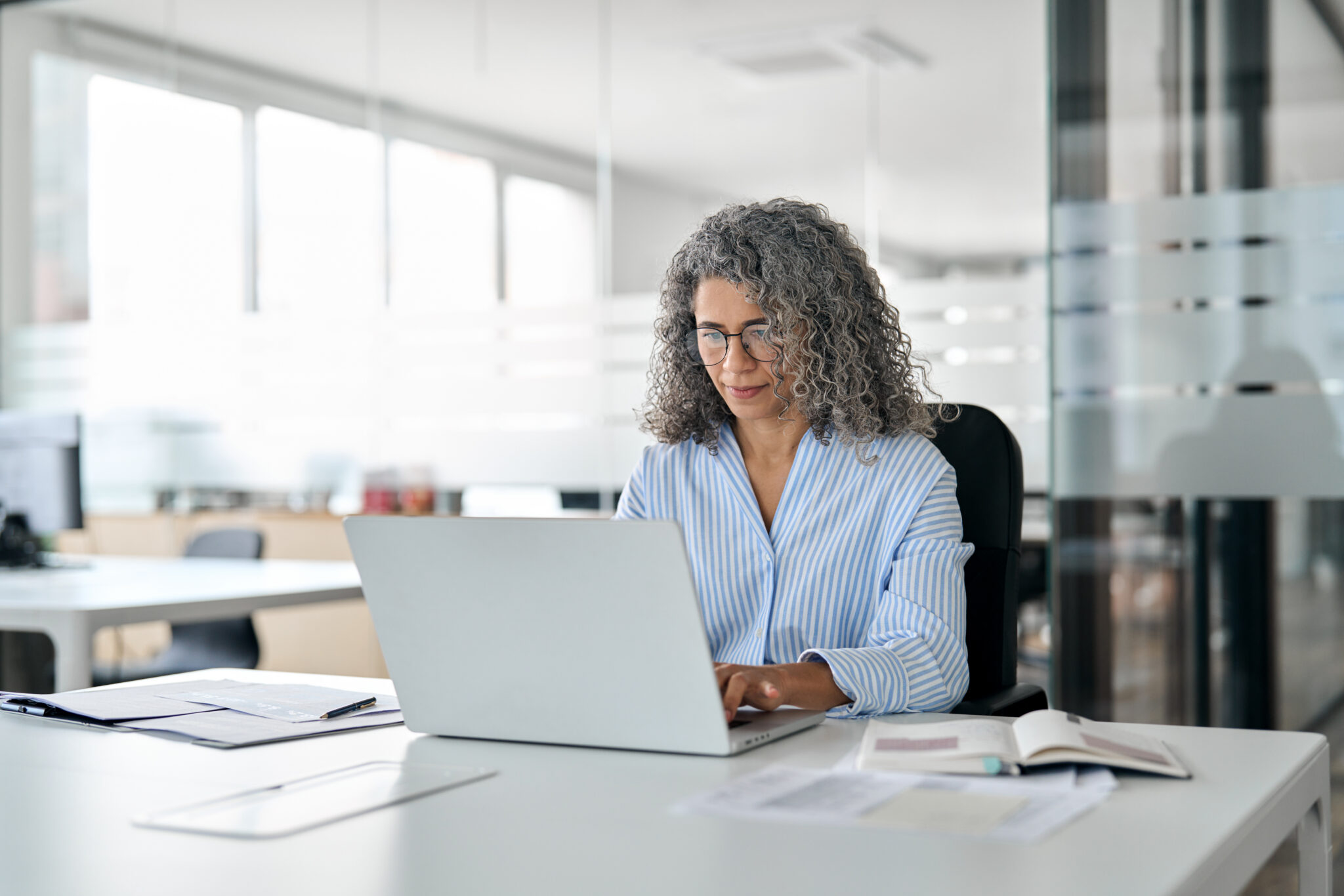 Woman with grey curly hair and blue striped shirt sitting at a desk typing on a laptop.