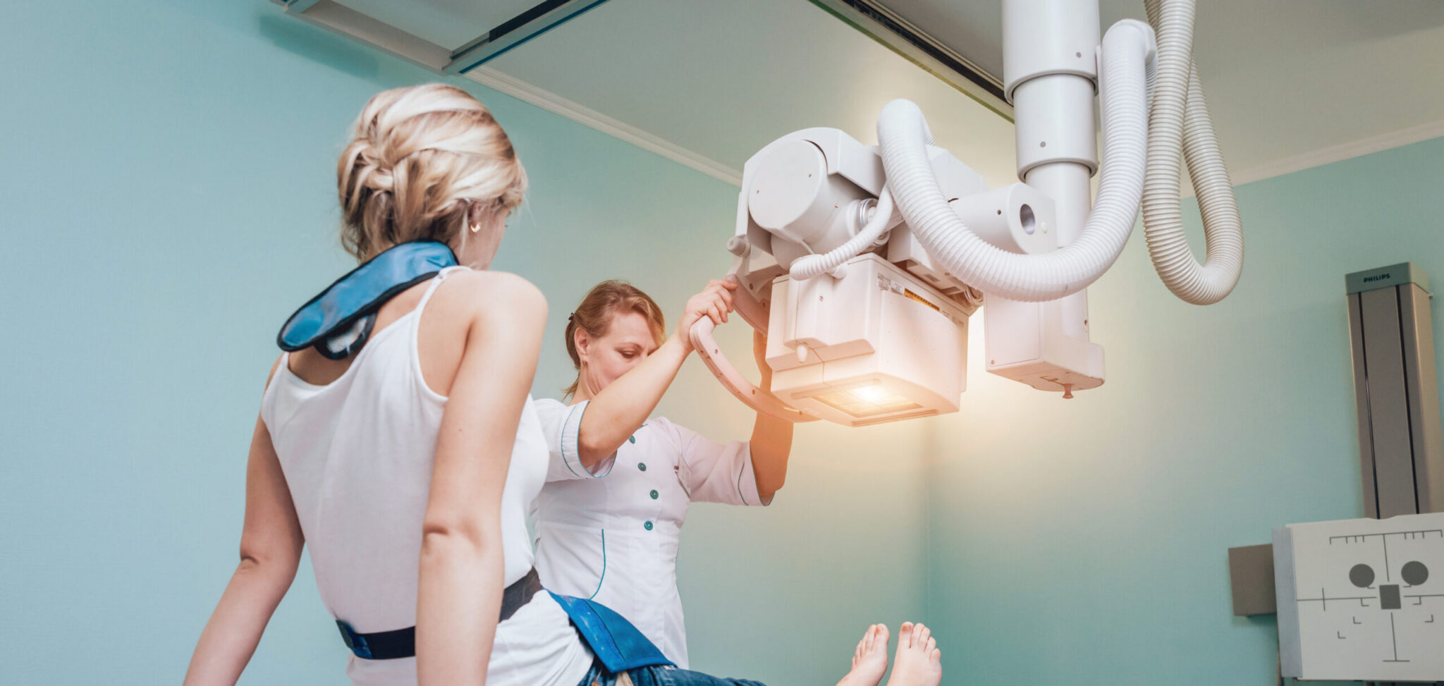 A radiologic technologist in white scrubs positions an overhead X-ray machine over a patient during an imaging exam in a light blue clinical room.