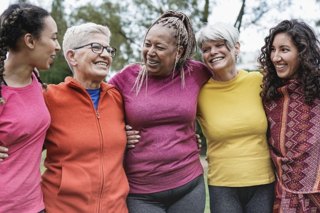 Five women of varying ages standing arm in arm, smiling outdoors.