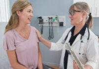Two shot of woman talking to her reassuring primary care doctor in exam room. Middle aged patient having appointment with female senior physician