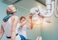 A radiologic technologist in white scrubs positions an overhead X-ray machine over a patient during an imaging exam in a light blue clinical room.