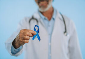 A doctor in a white coat with a stethoscope holds up a blue awareness ribbon toward the camera against a light blue background.