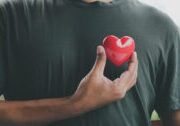 Person holding a small red heart to their chest outdoors.