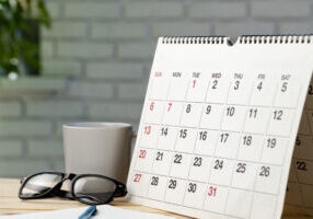 Calendar on a desk next to a pair of glasses, a mug, and a blue pen.