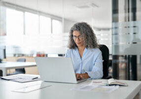 Woman with grey curly hair and blue striped shirt sitting at a desk typing on a laptop.
