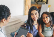Young family sitting on sofa listening to the advice from a female doctor using digital tablet. Doctor on house call giving medical consultation to a young family.