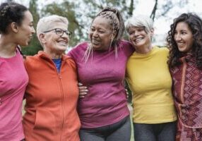 Five women of varying ages standing arm in arm, smiling outdoors.
